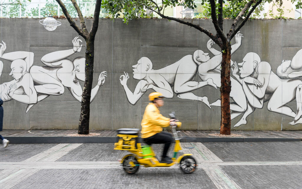 Gelb gekleideter Rollerfahrer fährt in Shenzhen an mit Graffitikunst besprühter Betonwand vorbei