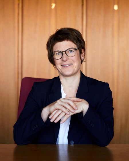 Veronika Grimm im Hosenanzug vor einer Holzwand sitzend