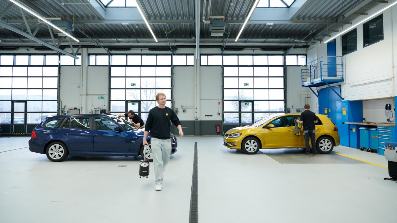 Drei Mechaniker arbeiten in einer hellen Werkstatthalle an einem blauen und einem gelben Kompaktwagen Drei Mechaniker arbeiten in einer hellen Werkstatthalle an einem blauen und einem gelben Kompaktwagen