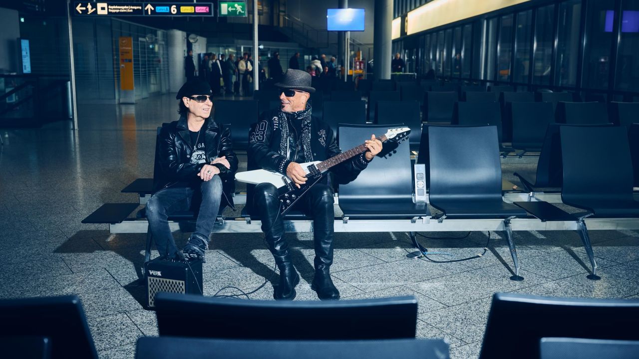 Zwei Bandmitglieder der Scorpions sitzen in einem Wartebereich am Flughafen, einer hält eine E-Gitarre Zwei Bandmitglieder der Scorpions sitzen in einem Wartebereich am Flughafen, einer hält eine E-Gitarre