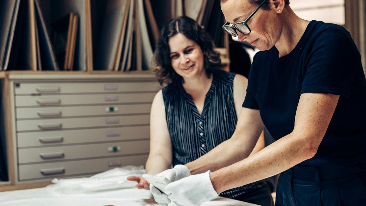 A restorer wearing white gloves stands at a work table holding a piece of porcelain in her hands, while a second woman watches with a smile. A restorer wearing white gloves stands at a work table holding a piece of porcelain in her hands, while a second woman watches with a smile.