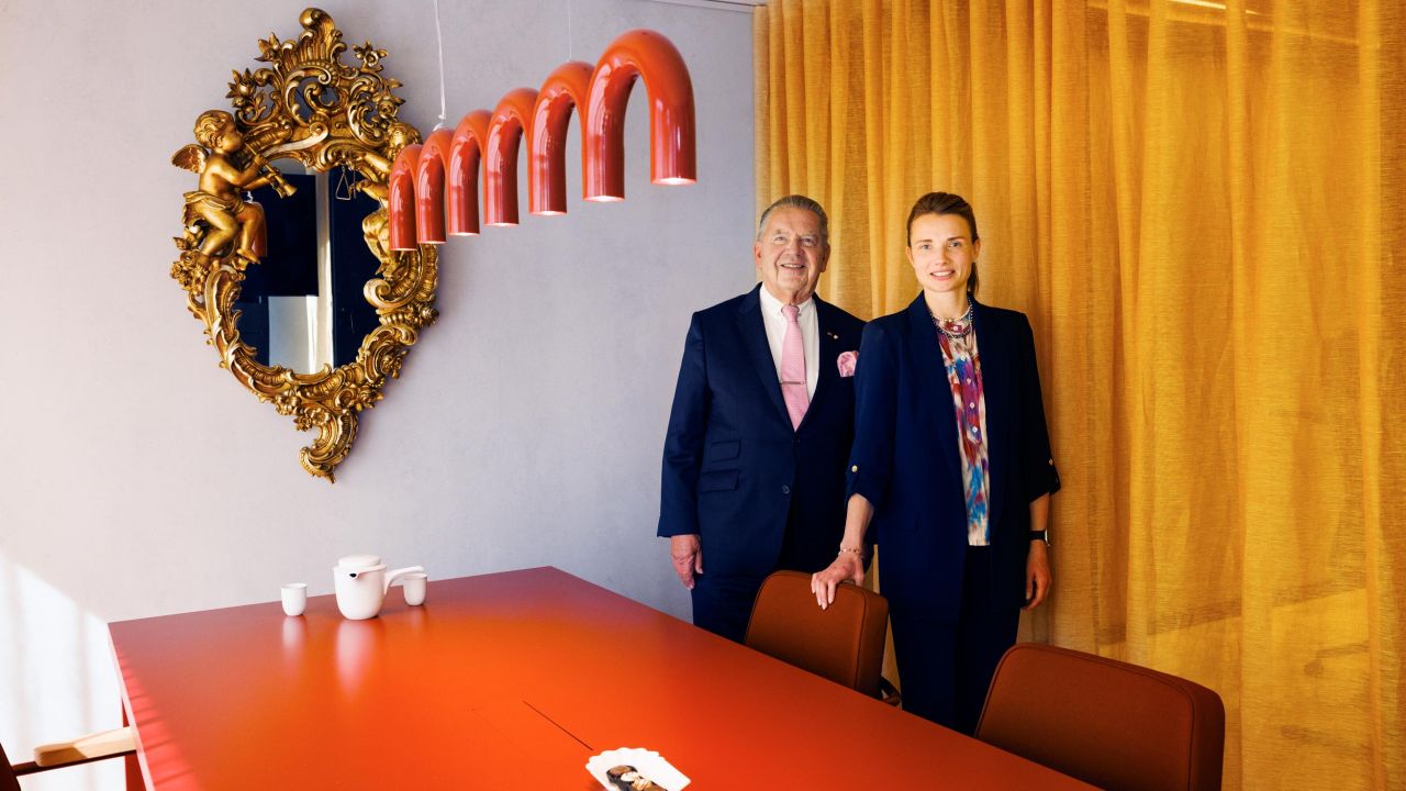 A smiling man and woman pose in a modern conference room in front of an orange table and yellow curtains A smiling man and woman pose in a modern conference room in front of an orange table and yellow curtains