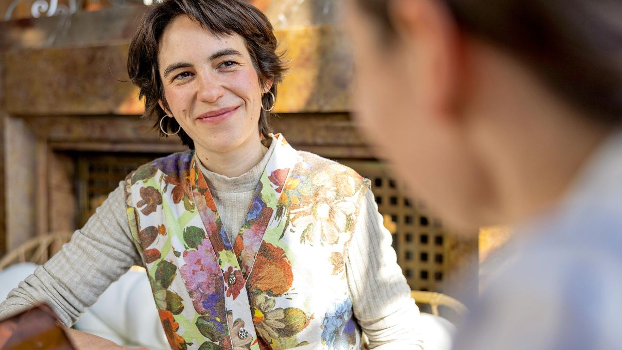 A smiling woman with short hair and a patterned vest is sitting at a table and looking at another person. A smiling woman with short hair and a patterned vest is sitting at a table and looking at another person.