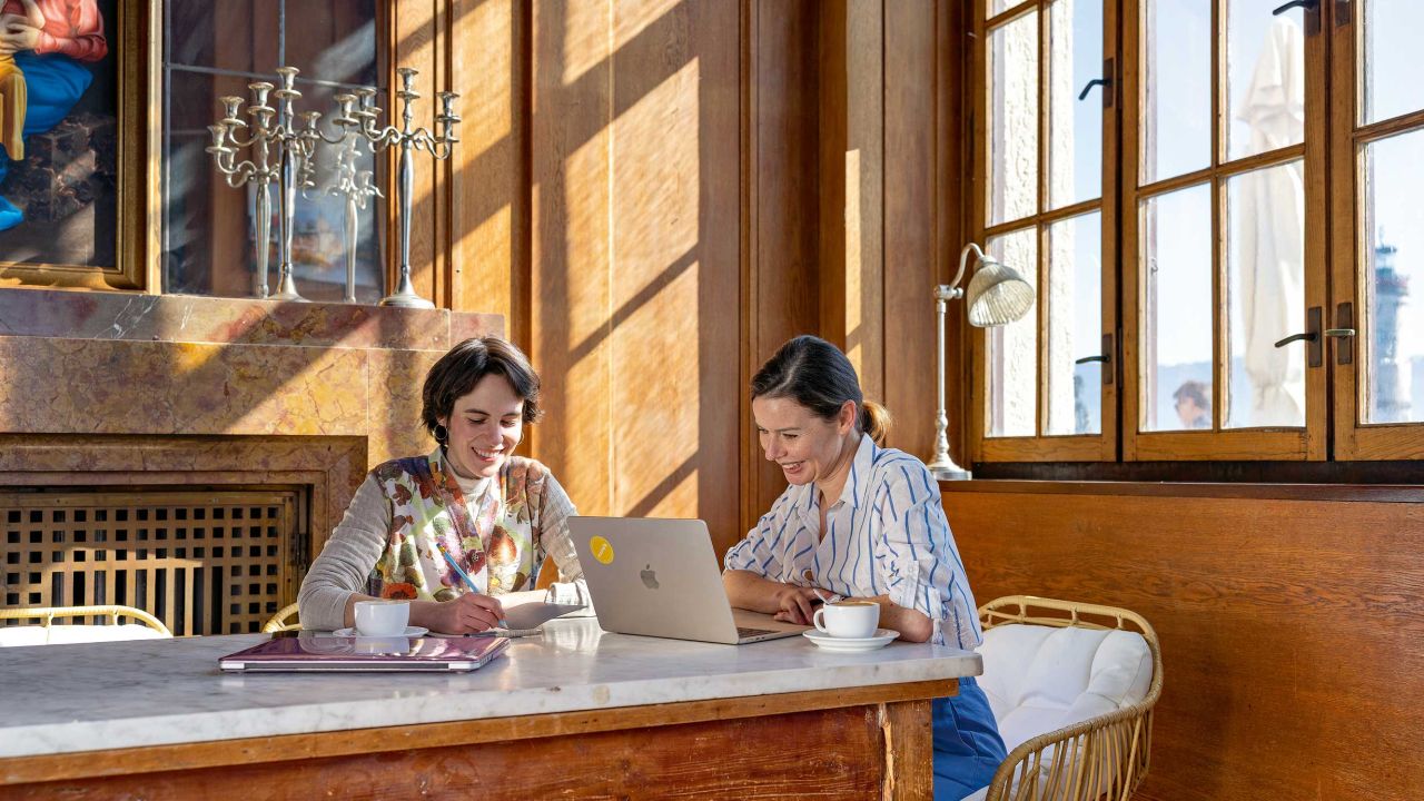 Two women are sitting with laptops at a wooden table in a bright room with wood paneling. Two women are sitting with laptops at a wooden table in a bright room with wood paneling.