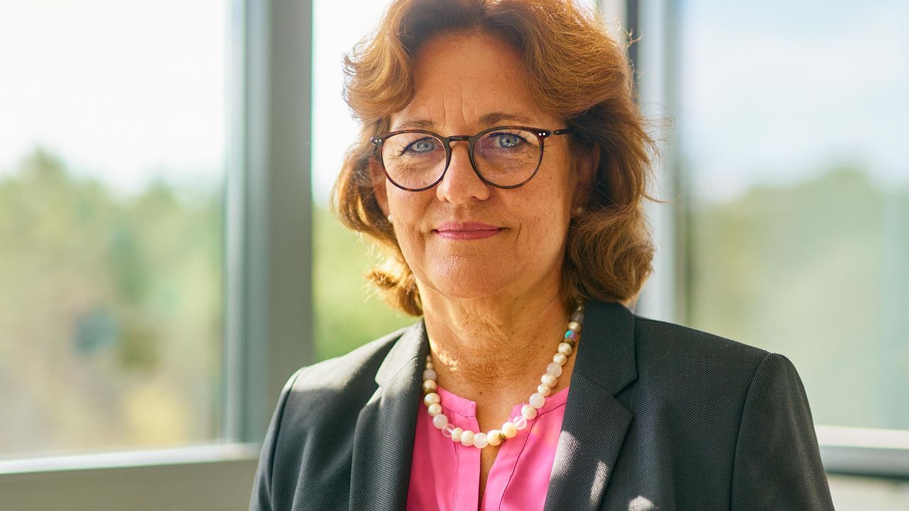 Close-up of a smiling woman in business attire with glasses and a pearl necklace in front of a large window Close-up of a smiling woman in business attire with glasses and a pearl necklace in front of a large window