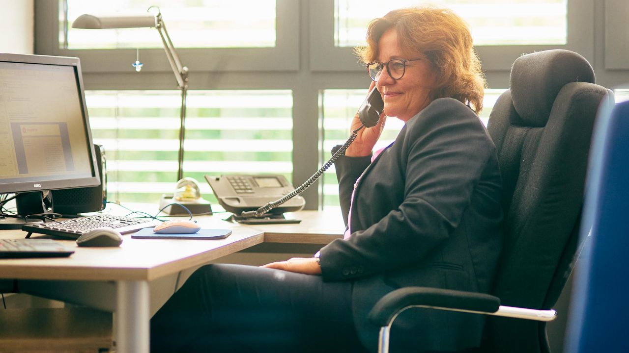 A woman in a dark business suit is smiling while talking on the phone at her desk in her office A woman in a dark business suit is smiling while talking on the phone at her desk in her office