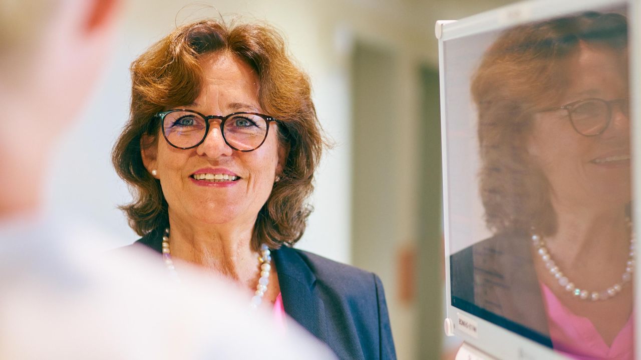 Smiling woman with glasses and a pearl necklace in conversation, her reflection is visible on a monitor in the foreground Smiling woman with glasses and a pearl necklace in conversation, her reflection is visible on a monitor in the foreground