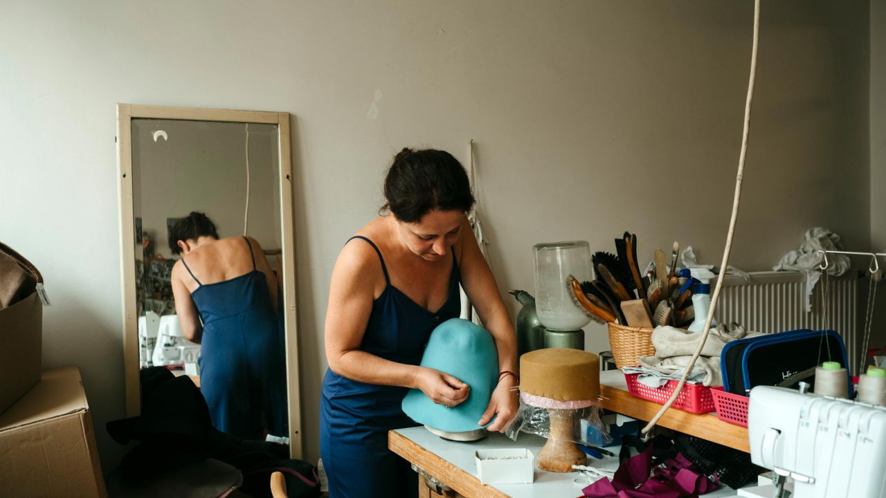 A milliner works on a light blue hat at a workbench, with her reflection visible in the mirror behind her. A milliner works on a light blue hat at a workbench, with her reflection visible in the mirror behind her.