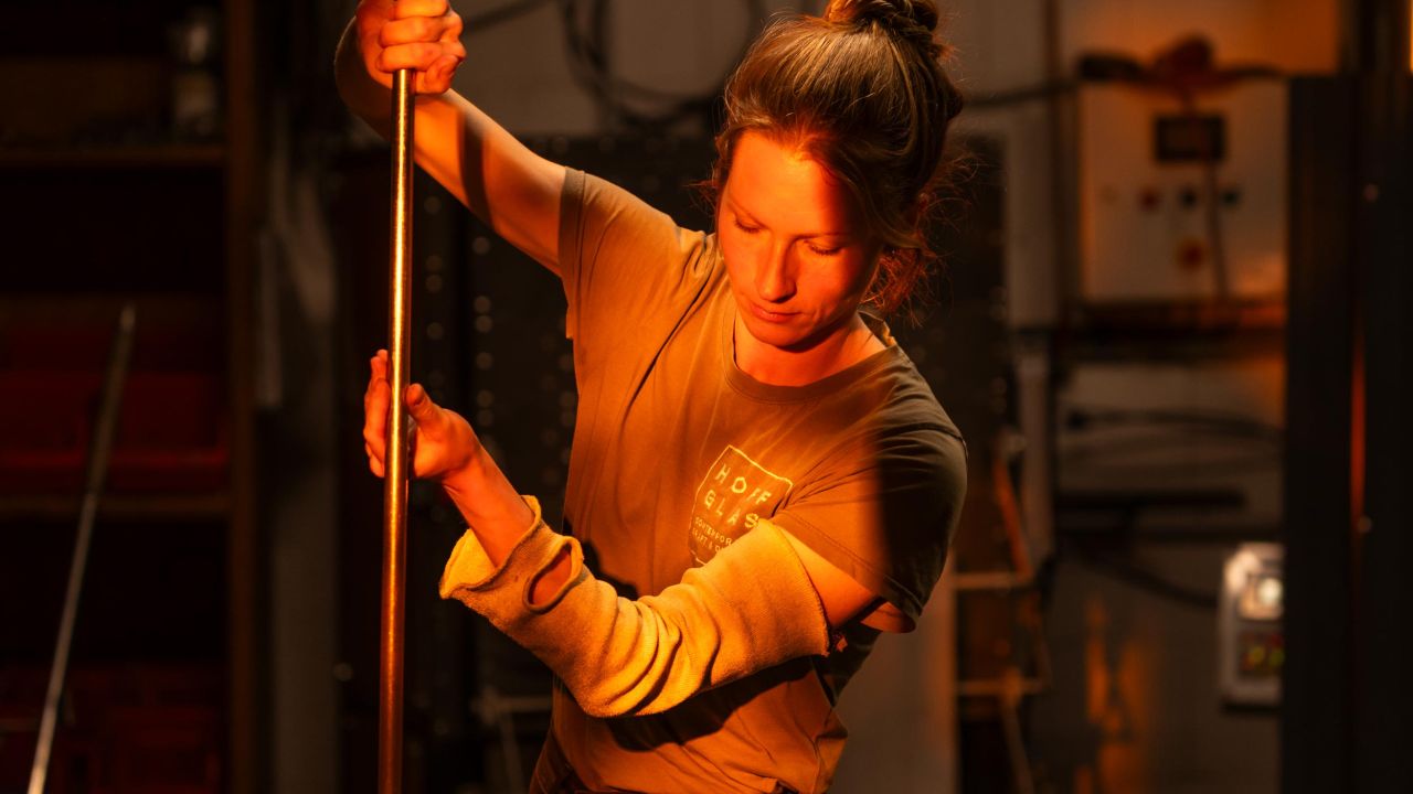 A glassblower dressed in dark clothing holds a long, vertical glassblower's pipe in her hands in a warmly lit workshop. A glassblower dressed in dark clothing holds a long, vertical glassblower's pipe in her hands in a warmly lit workshop.