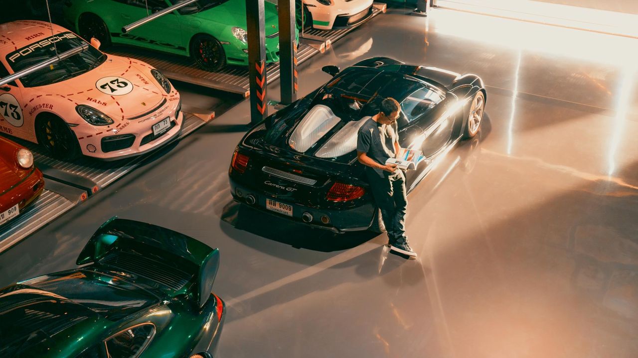 Man leaning against a Carrera GT in a garage, leafing through a magazine, surrounded by several parked Porsche sports cars Man leaning against a Carrera GT in a garage, leafing through a magazine, surrounded by several parked Porsche sports cars