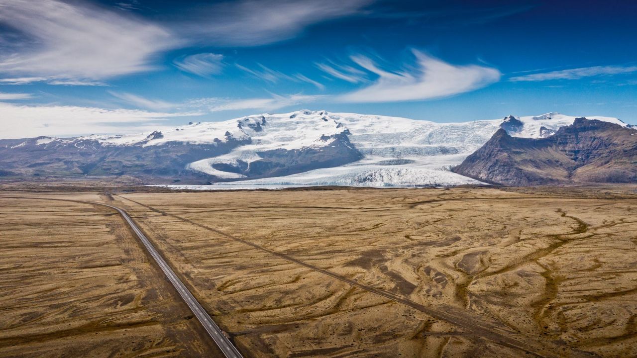 Aerial view of an empty road leading through a barren plain with glaciers and mountains in Iceland. Aerial view of an empty road leading through a barren plain with glaciers and mountains in Iceland.