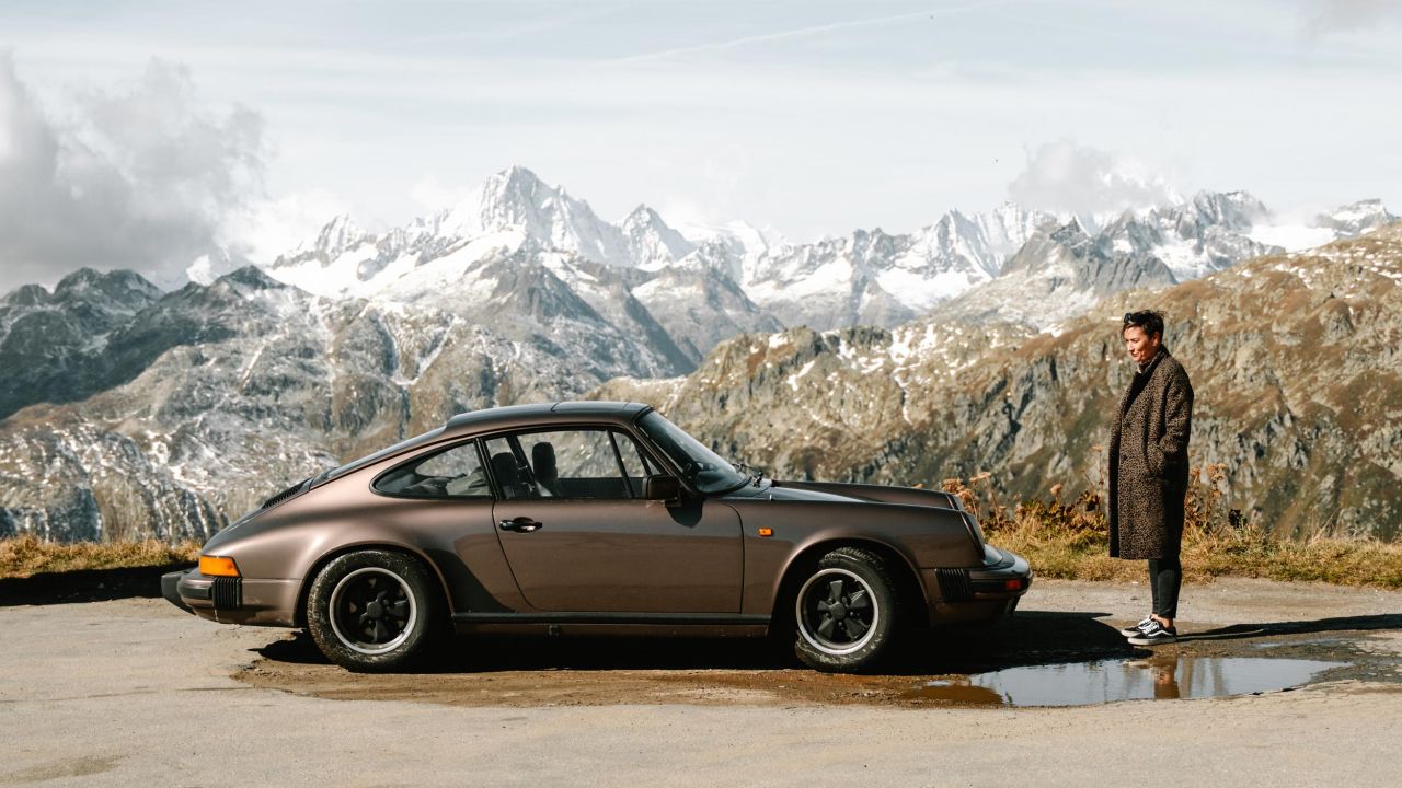 Ein brauner Porsche 911 steht auf einer Bergstraße vor einem Panorama schneebedeckter Alpengipfel, neben dem Auto steht eine Frau Ein brauner Porsche 911 steht auf einer Bergstraße vor einem Panorama schneebedeckter Alpengipfel, neben dem Auto steht eine Frau