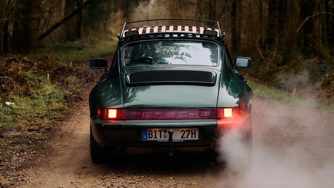 Heckansicht eines dunkelgrünen Porsche 911 mit Dachgepäckträger, der auf einem Waldweg fährt Heckansicht eines dunkelgrünen Porsche 911 mit Dachgepäckträger, der auf einem Waldweg fährt