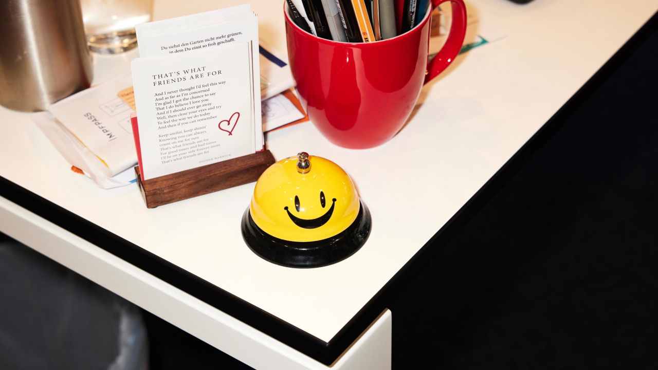 Gelb-schwarze Tischglocke mit einem Smiley steht auf einem weißen Schreibtisch neben einer Visitenkartenhalterung und einer roten Stiftekiste Gelb-schwarze Tischglocke mit einem Smiley steht auf einem weißen Schreibtisch neben einer Visitenkartenhalterung und einer roten Stiftekiste