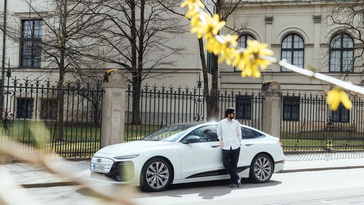 A man is leaning against the side of a white Audi A6 Sportback e-tron parked in an urban setting.