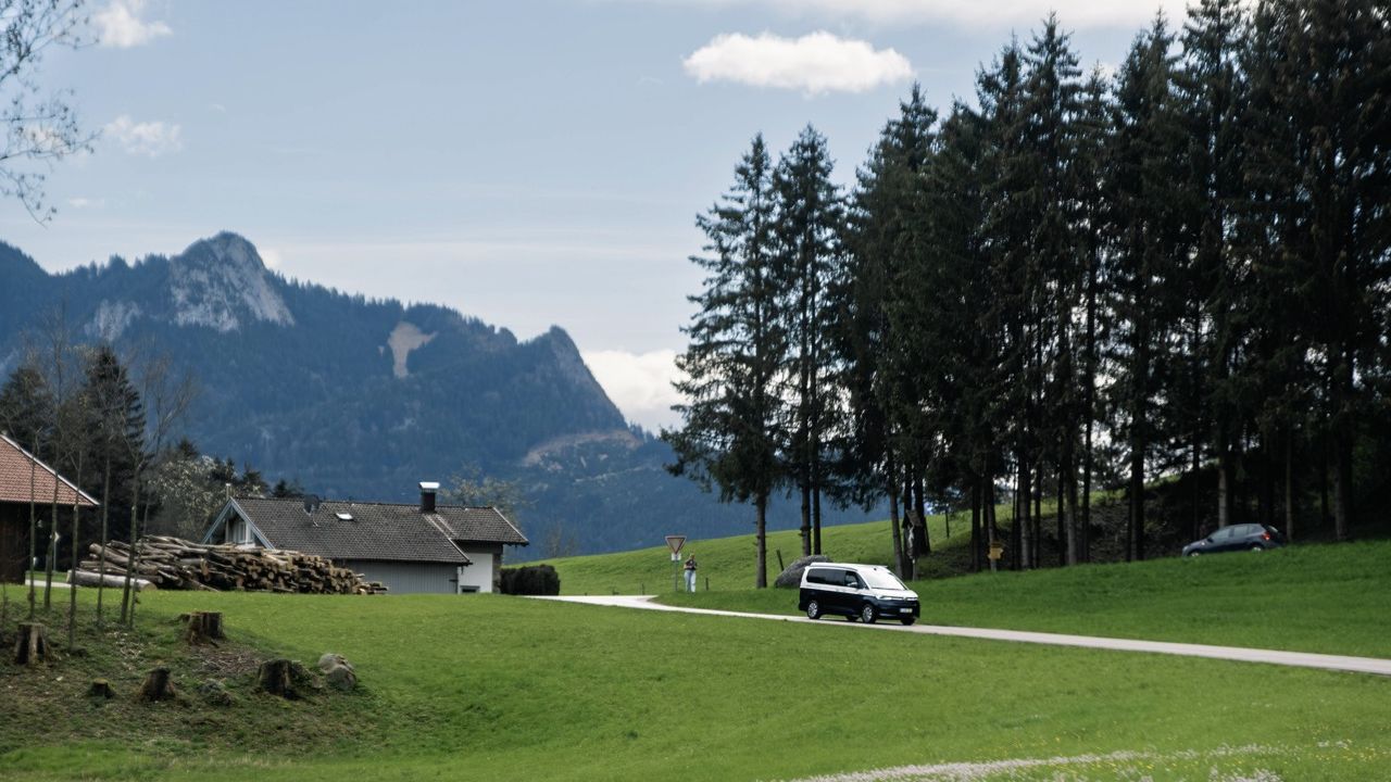 Dark VW California camper van driving on a narrow road through a green hilly landscape in the foothills of the Alps.